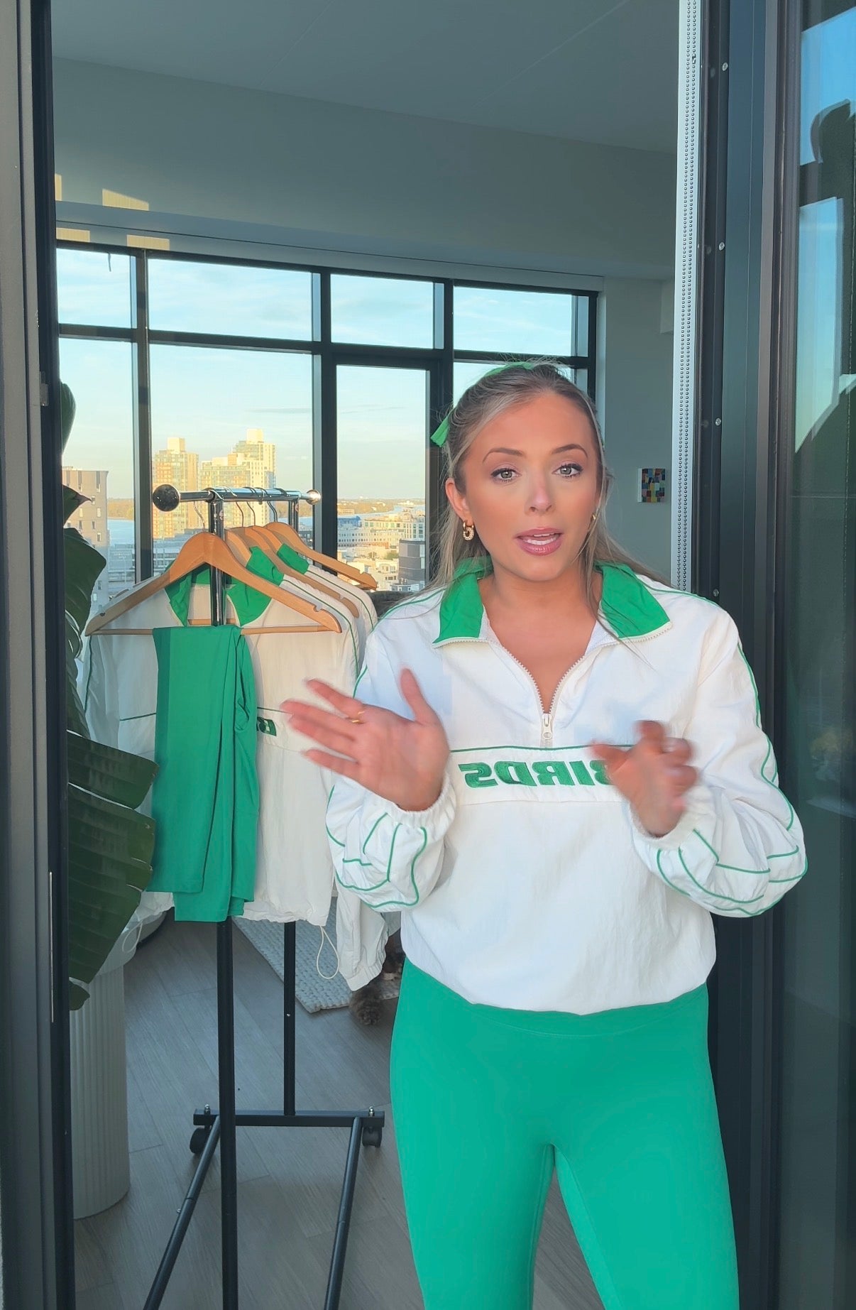 South & Green Co-Founder and Designer, Brooke, wearing the South & Green BIRDS Windbreaker and Kelly Green Yoga Pants  while standing in a room with a cityscape view.