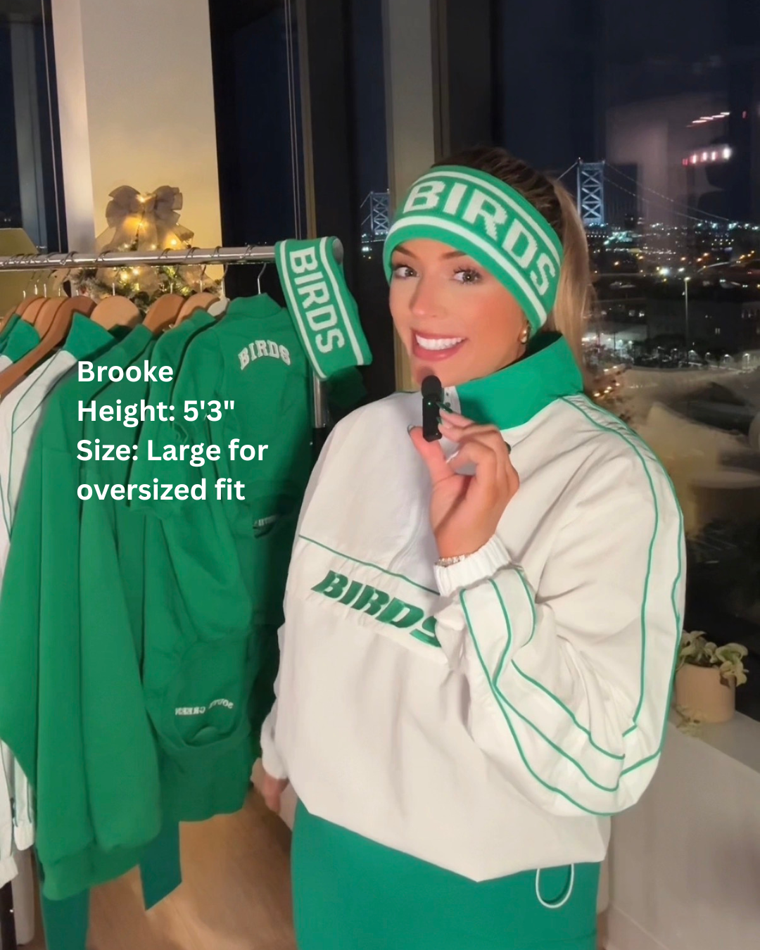 Brooke wearing South & Green's BIRDS Windbreaker, Kelly Green Yoga Pants, and BIRDS Ski Headband, standing in a room with cityscape view.